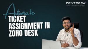 A Zentegra consultant sitting with a laptop and pointing upward beside text that reads “Automate Ticket Assignment in Zoho Desk” on a dark blue business-themed background.